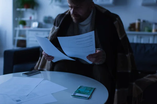 Hombre bajo una manta caliente mirando las facturas de pago cerca del dinero y el teléfono inteligente durante el corte de electricidad - foto de stock