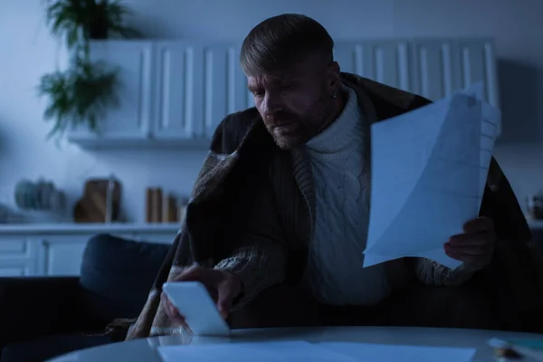 Hombre preocupado sosteniendo facturas de pago y mirando el teléfono celular durante el apagón de energía - foto de stock