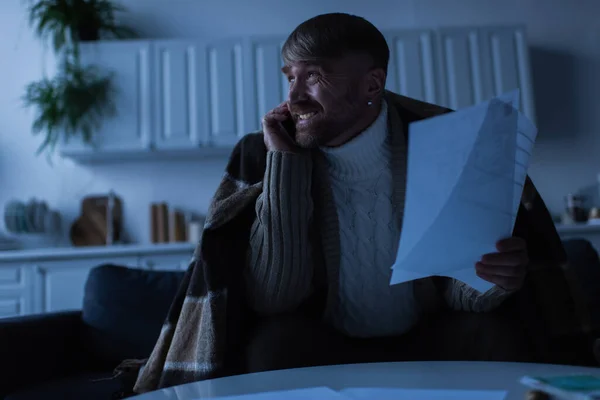 Нервовий і заморожений чоловік розмовляє по смартфону, тримаючи рахунки в сутінках — стокове фото