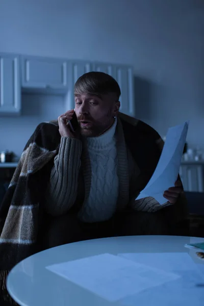 Shocked man under warm blanket holding invoices and talking on smartphone during power outage — Stock Photo