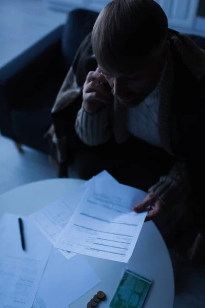 Hombre congelado sosteniendo facturas de pago y hablando en el teléfono móvil durante el apagón de energía en casa - foto de stock