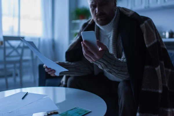 Frozen man with invoices looking at smartphone near money on table in twilight — Stock Photo