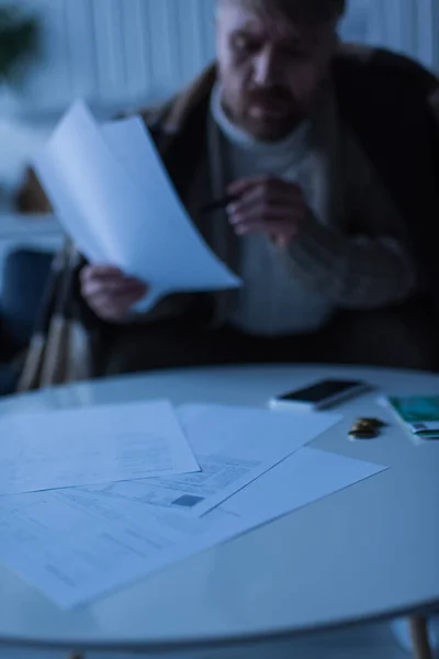Hombre borroso mirando las facturas cerca de la mesa con dinero y teléfono inteligente en el crepúsculo - foto de stock