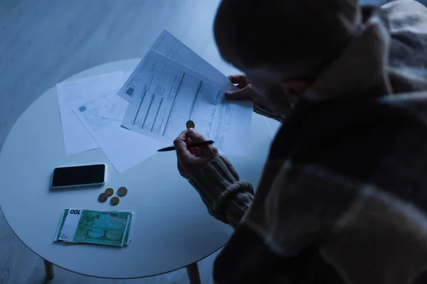 Vista de ángulo alto del hombre sosteniendo facturas cerca del dinero y el teléfono móvil en el crepúsculo - foto de stock