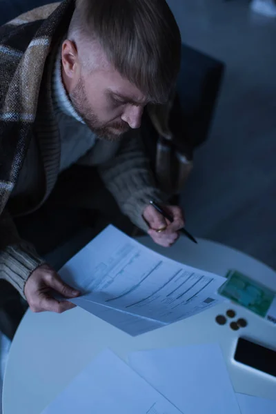High angle view of man in warm blanket looking at invoices near blurred money during energy blackout — Stock Photo