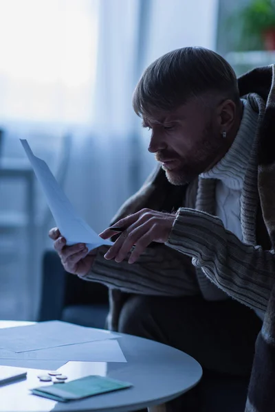 Hombre congelado en suéter caliente y manta mirando las facturas de pago durante el apagado de la electricidad - foto de stock