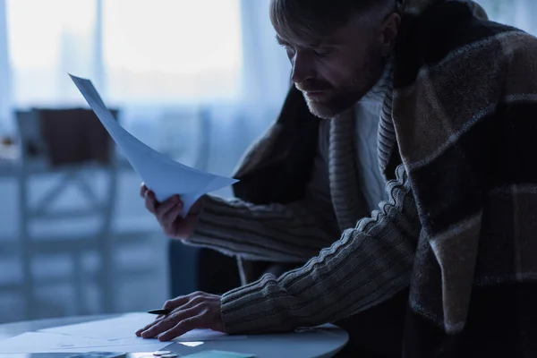 Homme congelé recouvert d'une couverture chaude regardant les factures de paiement au crépuscule — Photo de stock
