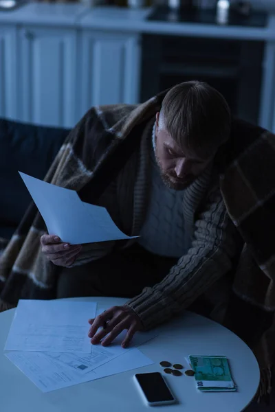 Человек сидит под теплым одеялом и смотрит на счета рядом с деньгами во время отключения энергии — стоковое фото