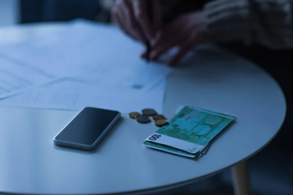 Billetes y monedas en euros cerca de smartphone con pantalla en blanco y hombre recortado con facturas de pago en un fondo borroso - foto de stock