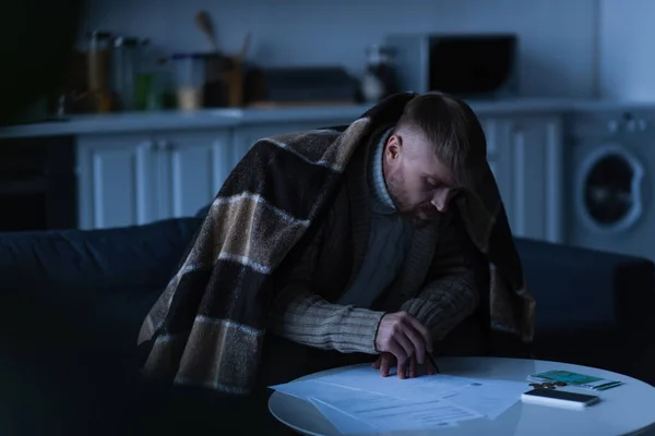 Man sitting under blanket and looking at payment bills during energy blackout — Stock Photo
