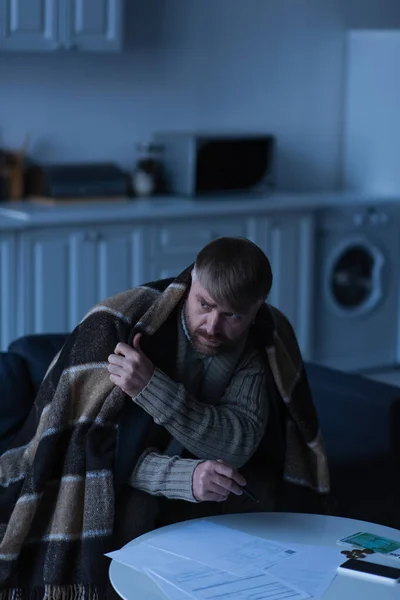 Расстроенный и замороженный человек сидит под теплым одеялом возле счетов и денег в сумерках — стоковое фото
