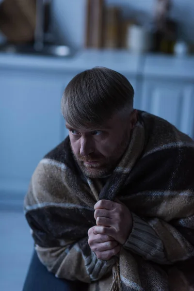 Unzufriedener und erstarrter Mann, der bei Stromausfall unter einer warmen Decke sitzt und wegschaut — Stockfoto