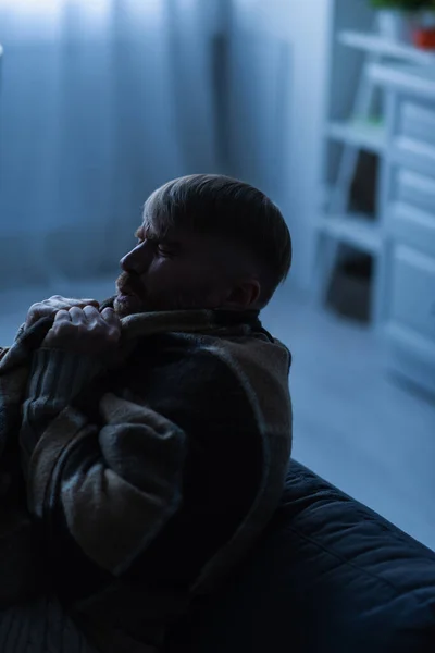 Hombre congelado con los ojos cerrados sentado en casa bajo una manta caliente durante el apagón eléctrico - foto de stock