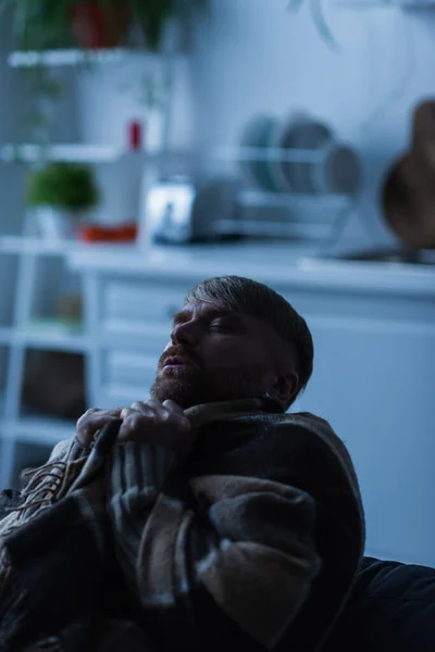 Homem congelado envolto em cobertor quente sentado na cozinha borrada com olhos fechados — Fotografia de Stock