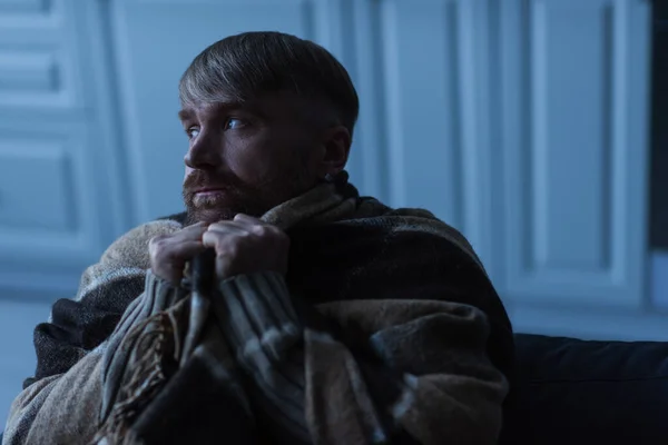 Upset and frozen man looking away while sitting under warm blanket during energy shutdown — Stock Photo