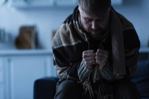 Hombre deprimido con la cabeza inclinada sentado debajo de una manta caliente en casa durante el apagón de energía - foto de stock