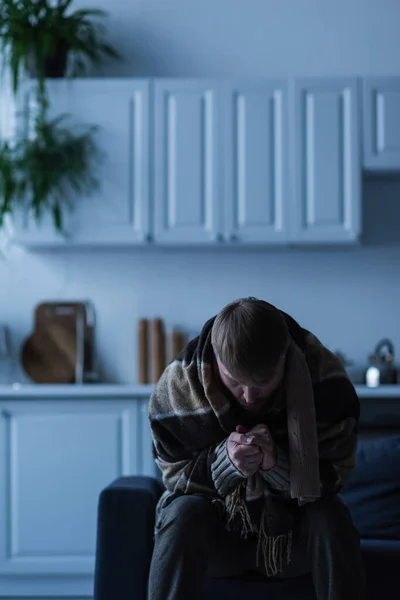 Hombre envuelto en las manos de calentamiento manta mientras está sentado en la cocina durante el apagado de energía - foto de stock