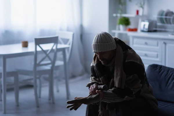 Frozen man in knitted hat sitting in kitchen under blanket and wearing warm gloves — Stock Photo