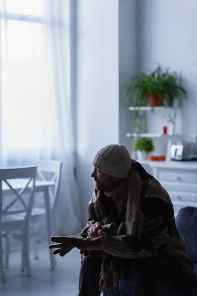 Frozen man in knitted hat wearing warm gloves while sitting under blanket and looking away in twilight — Stock Photo