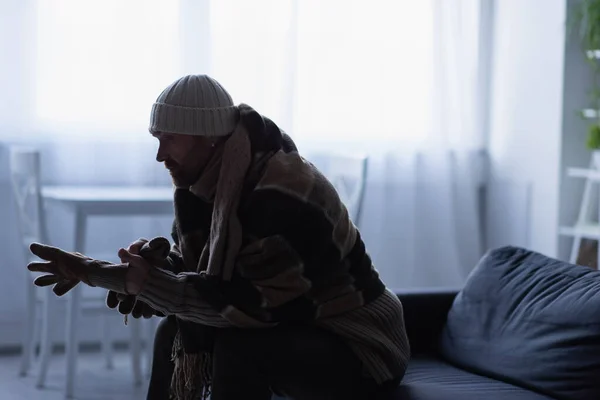 Side view of frozen man putting on gloves while sitting on couch in hat and warm blanket — Stock Photo