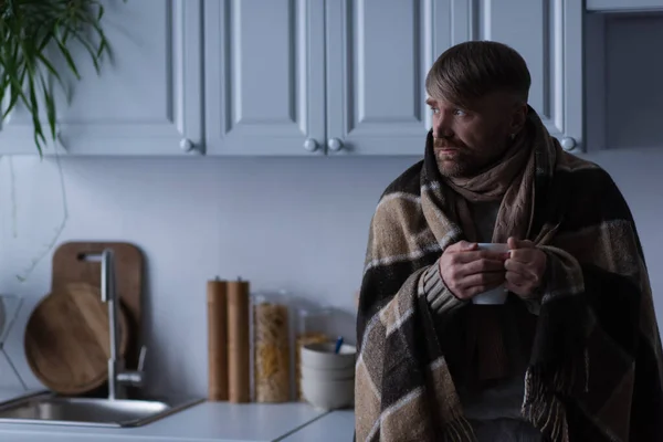 Homem congelado em cobertor quente segurando xícara de bebida quente e olhando para longe na cozinha — Fotografia de Stock