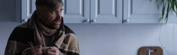 Замороженный человек в теплом одеяле, держа чашку горячего чая и глядя на дом в сумерках, баннер — стоковое фото