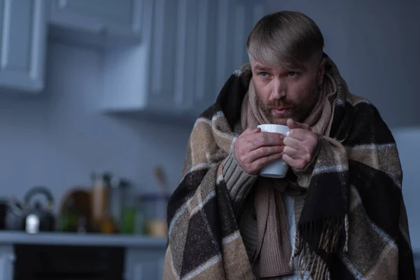 Man standing under warm blanket and blowing at hot tea during power outage at home — Stock Photo
