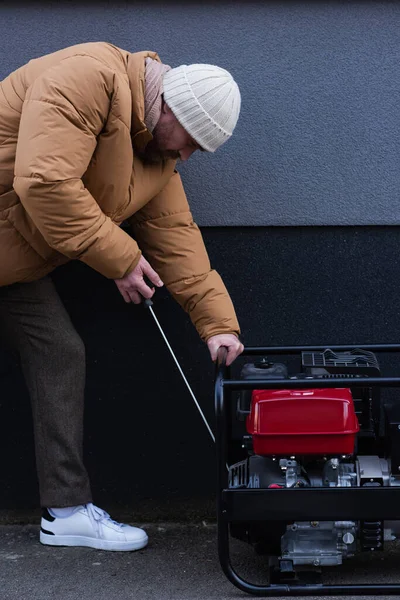 Vista lateral do homem em jaqueta e chapéu de malha começando gerador de eletricidade ao ar livre — Fotografia de Stock