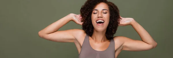 Positive Afroamerikanerin berührt lockiges Haar isoliert auf grünem Banner — Stockfoto