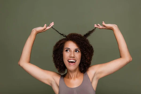 Positive african american woman touching curly hair and looking up isolated on green — Stock Photo