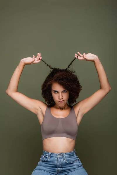Femme afro-américaine mécontente en haut et jeans touchant les cheveux bouclés et regardant la caméra isolée sur vert — Photo de stock