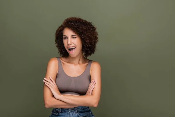 Pretty african american model crossing arms and winking at camera isolated on green — Stock Photo