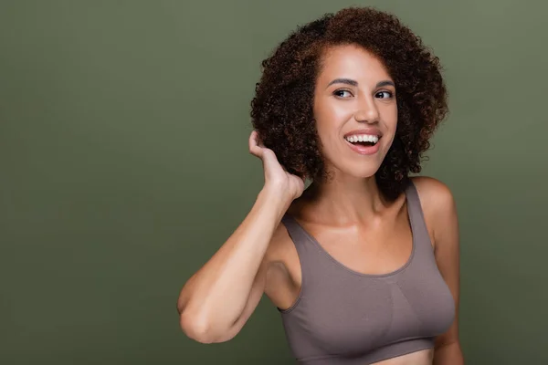 Sonriente mujer afroamericana en la parte superior tocando el pelo rizado y mirando hacia otro lado aislado en verde - foto de stock