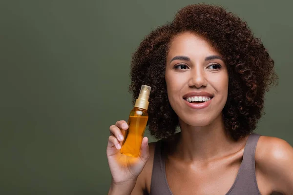 Femme afro-américaine bouclée tenant de l'huile cosmétique pour cheveux isolés sur vert — Photo de stock