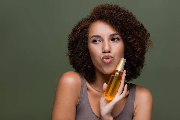 Mujer afroamericana joven sosteniendo aceite cosmético para el cabello rizado y labios pucheros aislados en verde — Stock Photo