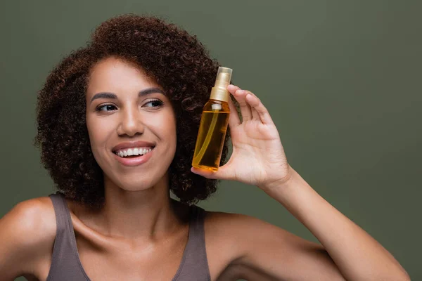 Sonriente mujer afroamericana mirando aceite de pelo cosmético aislado en verde - foto de stock