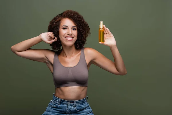 Curly african american woman in top and jeans holding cosmetic oil for hair isolated on green — Stock Photo