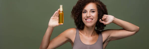 Bella donna afro-americana in alto guardando la fotocamera e tenendo olio per capelli isolato su verde, banner — Foto stock
