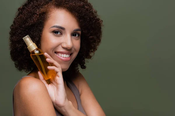 Retrato de una mujer afroamericana bastante rizada sosteniendo aceite cosmético y mirando a la cámara aislada en verde — Stock Photo