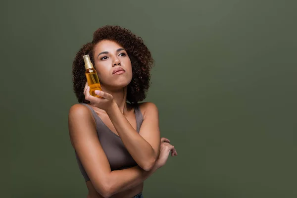 Young african american woman holding cosmetic oil and looking away isolated on green — Stock Photo