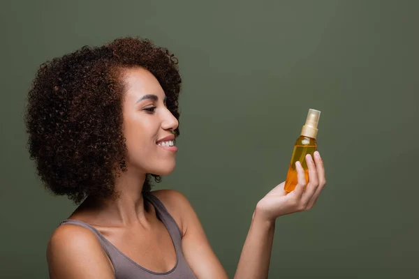 Vista lateral da mulher americana africana sorridente no topo olhando para o óleo cosmético isolado no verde — Fotografia de Stock