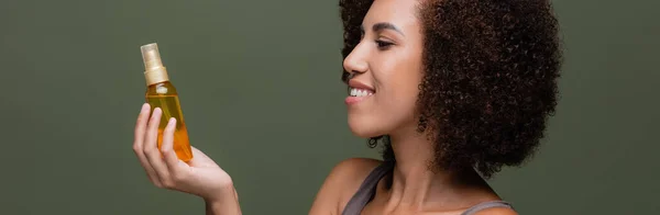 Vista laterale della donna riccia africana americana guardando l'olio cosmetico isolato sul verde, banner — Foto stock