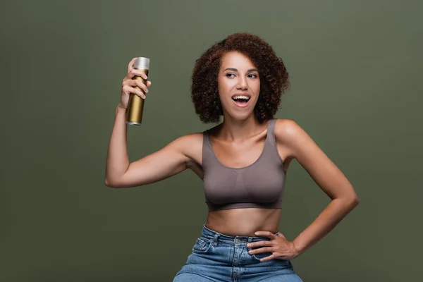 Cheerful african american woman in top holding hairspray isolated on green — Stock Photo