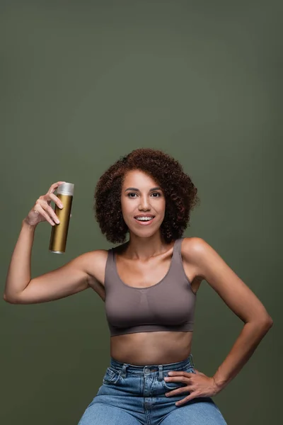 Pretty african american woman holding hairspray and looking at camera isolated on green — Stock Photo