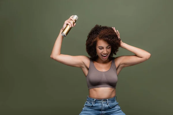 Cheerful african american woman in top and jeans applying hairspray isolated on green — Stock Photo
