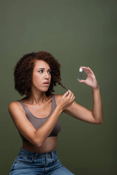 Preocupada mujer afroamericana tocando el pelo rizado y mirando aceite cosmético aislado en verde - foto de stock