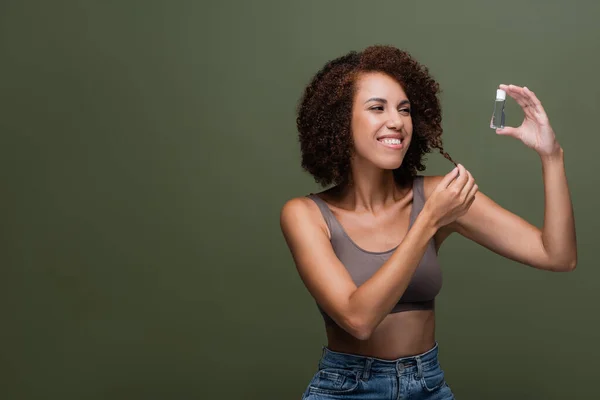 Positive african american woman holding cosmetic oil and touching curly hair isolated on green — Stock Photo