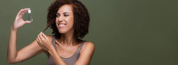 Веселая африканская американка, смотрящая на косметическое масло и трогательные кудрявые волосы, изолированные на зеленом, баннер — стоковое фото