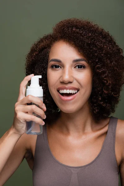 Porträt einer lächelnden Afroamerikanerin, die Kosmetikschaum in der Hand hält und isoliert auf Grün in die Kamera blickt — Stockfoto