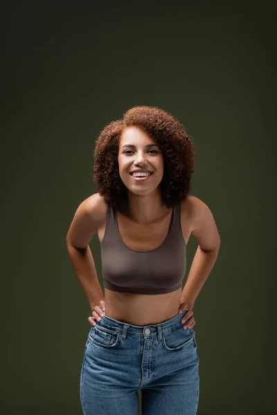Femme afro-américaine gaie en haut et jeans regardant la caméra isolée sur vert foncé — Photo de stock